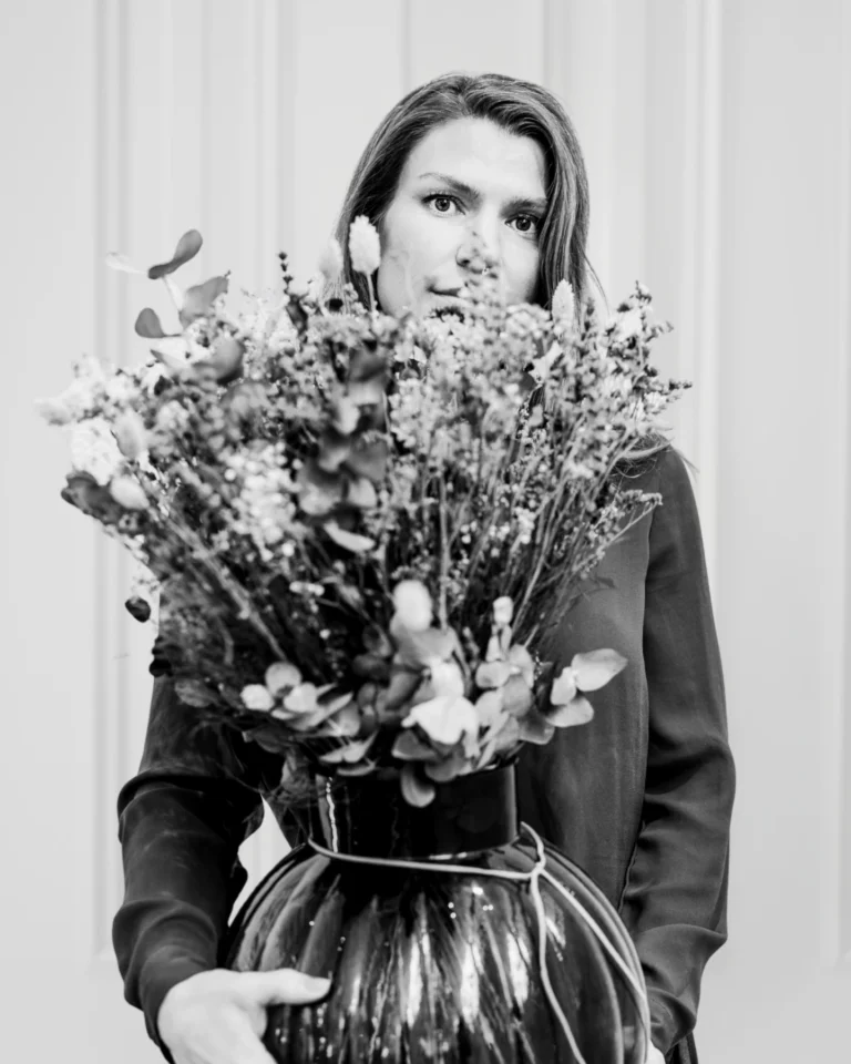 En kvinna står och håller i en stor vas fylld med diverse blommor och grönska, som delvis döljer hennes ansikte. Bilden är svartvit och har en enfärgad bakgrund.