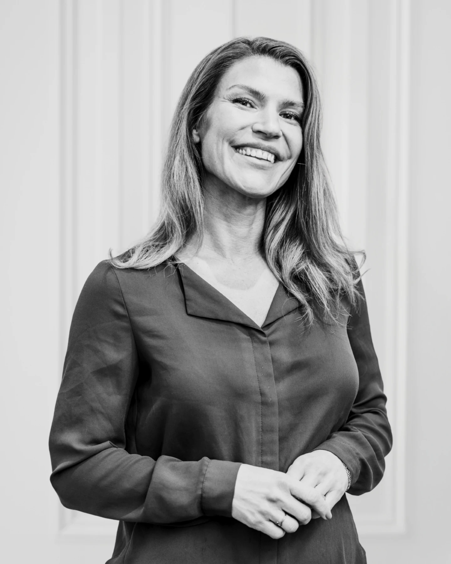 En kvinna med långt hår, klädd i en uppknäppt blus, står leende med händerna knäppta framför sig mot en ljus, panelklädd bakgrund. Fotot är i svartvitt.