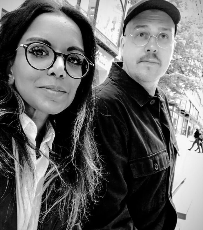 En kvinna och en man med glasögon och jacka poserar för en selfie på en gata i en stad. Bilden är i svartvitt och i bakgrunden syns byggnader och träd.