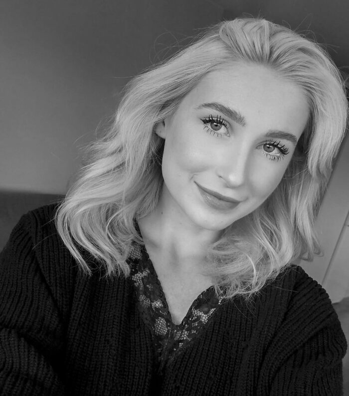 Svartvit selfie av en ung kvinna med vågigt, axellångt blont hår, klädd i en mörk stickad tröja över en spetstopp, som ler mjukt mot kameran.