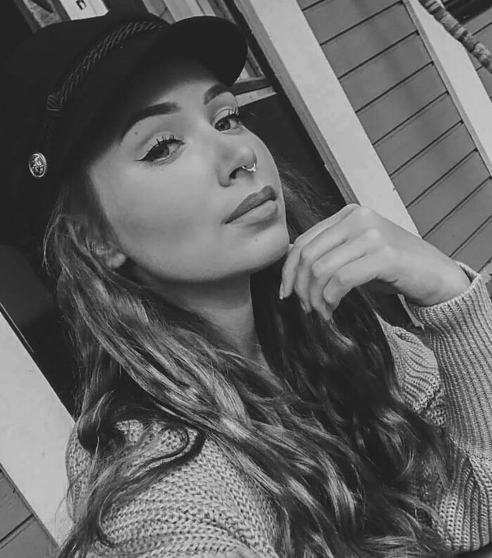 En ung kvinna med långt vågigt hår och en ring i näsan poserar för en selfie, iklädd keps och stickad tröja. Hon vilar hakan på sin hand och tittar självsäkert in i kameran. Bilden är i svartvitt.