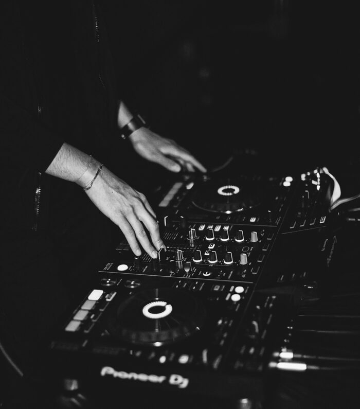 En persons händer justerar reglagen på en DJ-mixer och skivspelare, med olika knappar och vred upplysta, i en mörk, stämningsfull miljö.